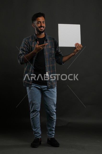 موك اب مربع أبيض، إعلانات وعروض تسويقية، استخدام لوحات توضيحية ووسائل تعليمية، بورتريه لشاب عربي خليجي سعودي مبتسم يرتدي الزي الكاجوال يمسك بيده لوحه بيضاء مربعه فارغة وينظر إلى الكاميرا، صورة الجسم بالكامل، خلفية سوداء