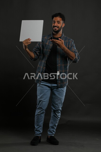 A white square mockup, advertisements and marketing presentations, the use of explanatory panels and teaching aids, a portrait of a smiling Gulf Arab Saudi man wearing a casual outfit, holding an empty square white board in his hand and looking at the camera, full body portrait, black background