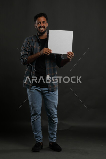 Gestures of joy looking at the camera, a white square mockup, advertisements and marketing presentations, the use of explanatory panels and teaching aids, a portrait of a smiling Gulf Arab Saudi man wearing a casual outfit holding a blank square white board in his hand, full body portrait, black background