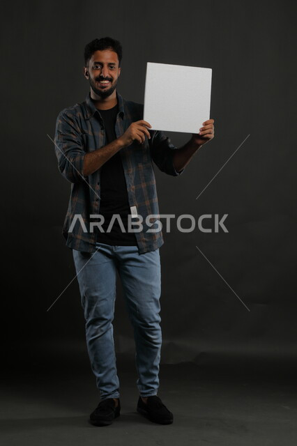 Advertisements and marketing presentations, a white square mockup, the use of explanatory panels and teaching aids, a portrait of a smiling Gulf Arab Saudi man wearing a casual outfit holding an empty square white board in his hand and looking at the camera, full body portrait, black background
