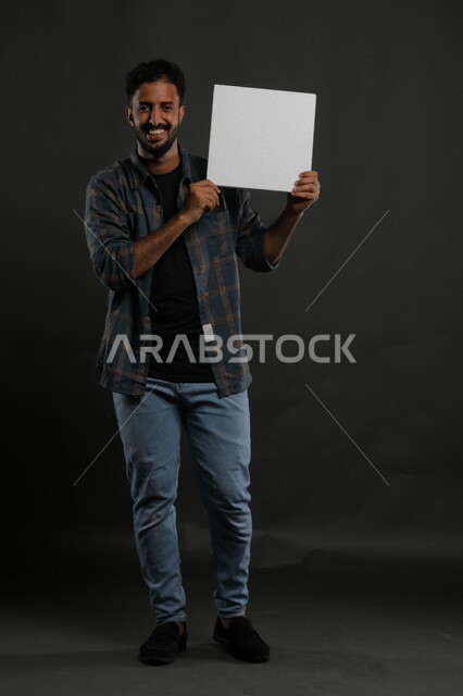عروضات وإعلانات تسويقية، موك اب مربع أبيض، استخدام لوحات توضيحية ووسائل تعليمية، بورتريه لشاب عربي خليجي سعودي مبتسم يرتدي الزي الكاجوال يمسك بيده لوحه بيضاء مربعه فارغة وينظر إلى الكاميرا، صورة الجسم بالكامل، خلفية سوداء