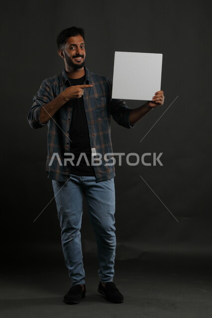الإشارة إلى لوحة بيضاء مربعة فارغة، إعلانات وعروض تسويقية، موك اب مربع أبيض، استخدام لوحات توضيحية ووسائل تعليمية، بورتريه لشاب عربي خليجي سعودي مبتسم يرتدي الزي الكاجوال ينظر إلى الكاميرا، صورة الجسم بالكامل، خلفية سوداء