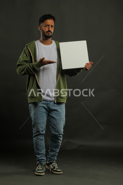 Gestures of anxiety and annoyance, advertisements and marketing presentations, white square mockup, portrait of a Saudi Gulf Arab man wearing casual clothes holding a blank square white board in his hand and looking at the camera, full body portrait, black background