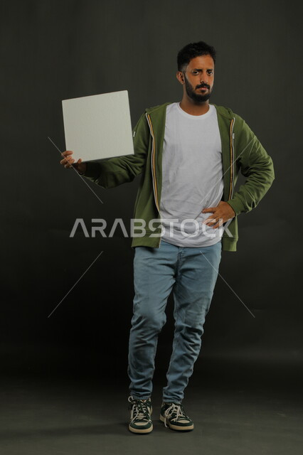الشعور بالقلق والانزعاج، الإعلانات والعروض التسويقية، موك اب مربع أبيض، بورتريه لرجل عربي خليجي سعودي يرتدي الزي الكاجوال يمسك بيده لوحه بيضاء مربعه فارغة وينظر إلى الكاميرا، صورة الجسم بالكامل، خلفية سوداء