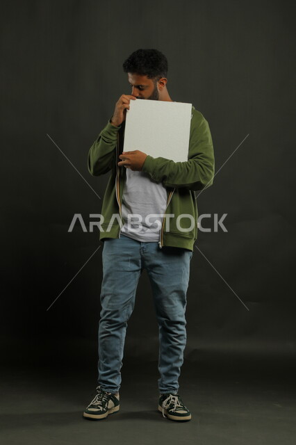 اعلانات وعروض تسويقية، موك اب مربع أبيض، استخدام لوحات توضيحية ووسائل تعليمية، بورتريه لشاب عربي خليجي سعودي مبتسم يرتدي الزي الكاجوال يمسك بيده لوحه بيضاء مربعه فارغة وينظر إلى الكاميرا، صورة الجسم بالكامل، خلفية سوداء