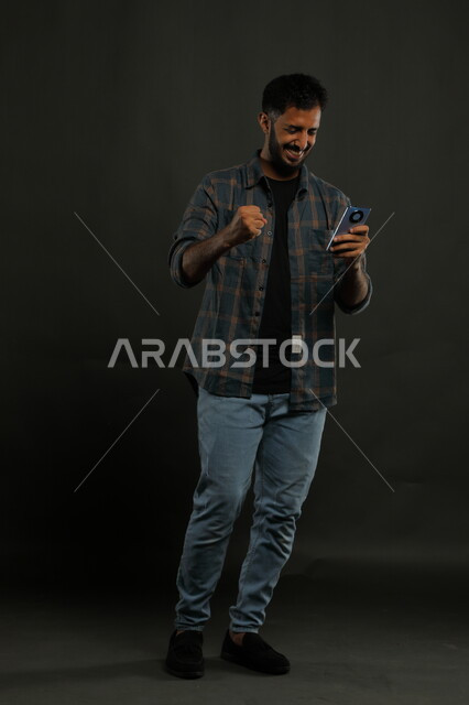 دور التقنية في توفير راحة أكبر للأفراد، الشعور بالحماس للنجاح وتحقيق الأهداف, بورتريه لشاب عربي سعودي خليجي يرتدي الزي الكاجوال يمسك الهاتف المحمول يرفع قبضته بإيماءات الفرح الشديد, السرور والفرح بالإنجازات, صورة الجسم بالكامل, خلفية سوداء.