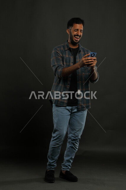 احتراف الوظائف التقنية وتكنولوجيا المعلومات، مشاهدة أخبار سارة على وسائل التواصل الاجتماعي، بورتريه لشاب عربي سعودي خليجي يرتدي اللباس الكاجوال يستخدم الهاتف الجوال بإيماءات الفرح والسعادة للفوز بالألعاب الالكترونية, صورة الجسم بالكامل, خلفية سوداء