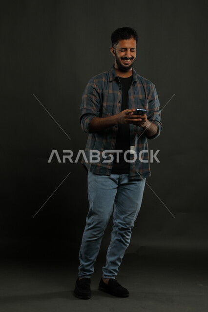 احتراف الوظائف التقنية وتكنولوجيا المعلومات، مشاهدة أخبار سارة على وسائل التواصل الاجتماعي، بورتريه لشاب عربي سعودي خليجي يرتدي اللباس الكاجوال يستخدم الهاتف الجوال بإيماءات الفرح والسعادة للفوز بالألعاب الالكترونية, صورة الجسم بالكامل, خلفية سوداء