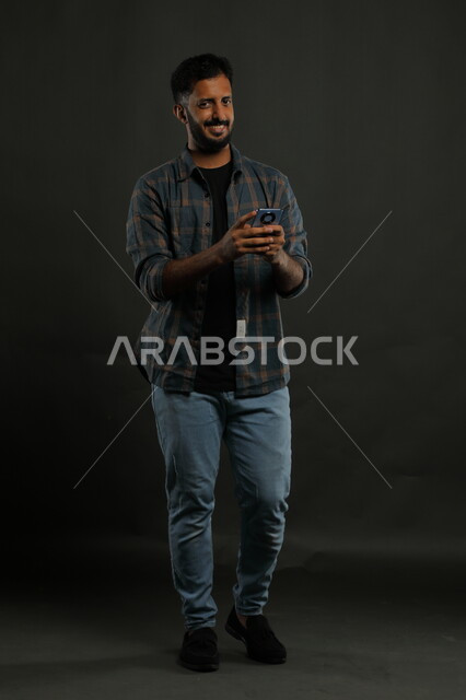 Looking at the camera with gestures of joy, pleasure and intelligence, professionalism in technical and information technology jobs, watching good news on social media, portrait of a smiling Saudi Arabian Gulf young man wearing casual clothes using a mobile phone, full body portrait, black background