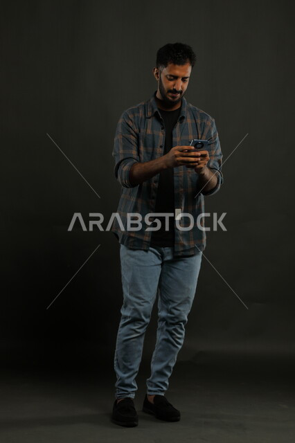 استخدام الأجهزة الحديثة في تطوير المهارات الشخصية، احتراف الوظائف التقنية وتكنولوجيا المعلومات، متابعة آخر أخبار السوشال ميديا، بورتريه لشاب عربي سعودي خليجي مبتسم يرتدي اللباس الكاجوال يستخدم الهاتف الجوال, صورة لكامل الجسم, خلفية سوداء