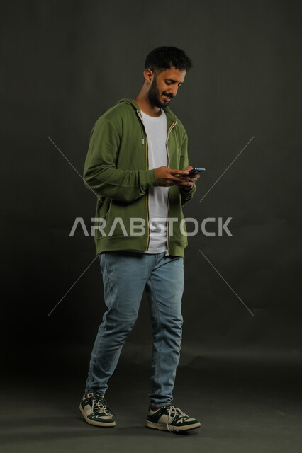 استخدام الأجهزة الحديثة في تطوير المهارات الشخصية، احتراف الوظائف التقنية وتكنولوجيا المعلومات، متابعة آخر أخبار السوشال ميديا، بورتريه لشاب عربي سعودي خليجي مبتسم يرتدي اللباس الكاجوال يستخدم الهاتف الجوال, صورة لكامل الجسم, خلفية سوداء