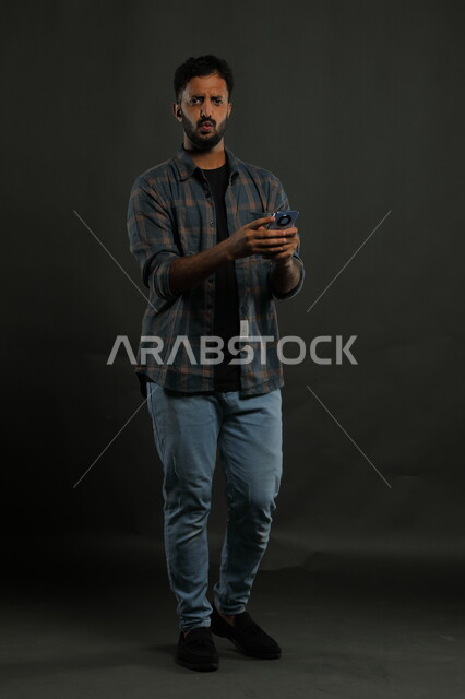خسارة المنتخب المفضل في المباراة، احتراف الوظائف التقنية وتكنولوجيا المعلومات، اللعب بالألعاب الاكترونية الهاتفية، بورتريه لشاب عربي سعودي خليجي يرتدي اللباس الكاجوال يستخدم الهاتف الجوال بإيماءات الدهشة والذهول, صورة لكامل الجسم, خلفية سوداء