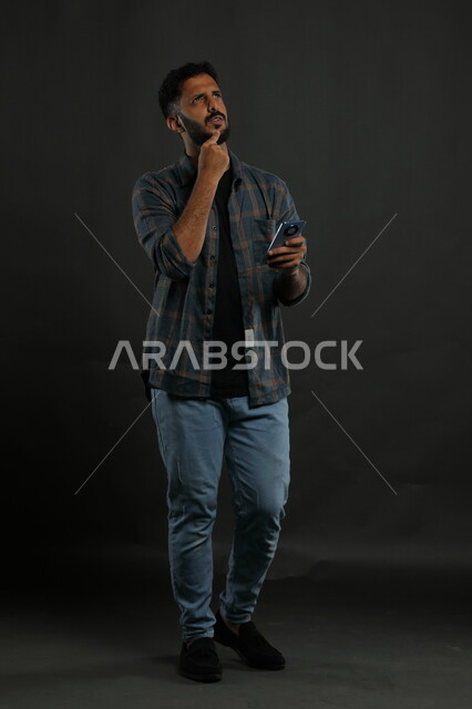 التأمل والتخطيط للمستقبل، بورتريه لشاب عربي خليجي سعودي يرتدي الزي الكاجوال يمسك بيده الهاتف المحمول يضع يده على ذقنه وينظر إلى الأعلى بإيماءات التفكيرلإيجاد حل لمشكلة ما باستخدام التكنولوجيا، صورة الجسم بالطول الكامل، خلفية سوداء