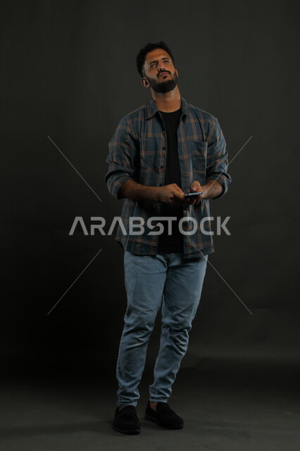 الشعور بالملل والتعب من ضغوطات العمل والحياة، بورتريه لشاب عربي خليجي سعودي يرتدي الزي الكاجوال يمسك بيده الهاتف المحمول ينظر إلى الأعلى بإيماءات التفكيرلإيجاد حل لمشكلة ما باستخدام التكنولوجيا، صورة الجسم بالطول الكامل، خلفية سوداء