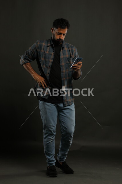 إنجاز الأعمال عن بعد باستخدام التكنولوجيا والتقنية الحديثة، الشعور بالملل والتعب من ضغوطات العمل والحياة، بورتريه لشاب عربي خليجي سعودي يرتدي الزي الكاجوال يمسك بيده الهاتف المحمول بإيماءات الانزعاج، صورة الجسم بالطول الكامل، خلفية سوداء