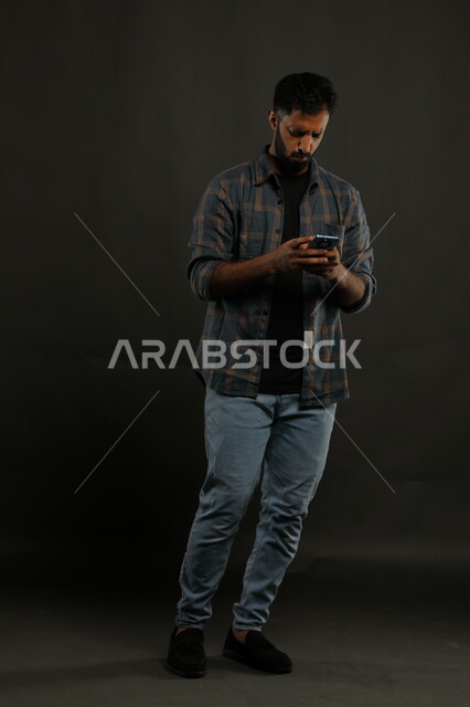إنجاز الأعمال عن بعد باستخدام التكنولوجيا والتقنية الحديثة، تلقي أخبار مزعجة من العمل، بورتريه لشاب عربي خليجي سعودي يرتدي الزي الكاجوال يمسك بيده الهاتف المحمول بإيماءات الانزعاج، صورة الجسم بالطول الكامل، خلفية سوداء