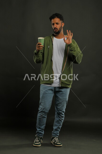 منتج محلي وطني، الحيوية والنشاط عند شرب الكافيين، إيماءات الإعجاب والاستمتاع بتناول المشروبات الساخنة، بورتريه لرجل عربي سعودي يرتدي الزي الكاجوال يحمل بيده كوب ورقي بإيماءات علامة جودة القهوة السعودية، صورة الجسم بالكامل، خليفية سوداء