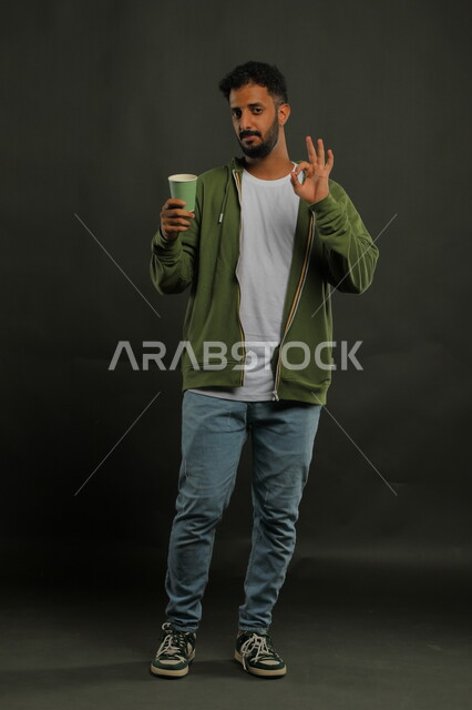 منتج محلي وطني، الحيوية والنشاط عند شرب الكافيين، إيماءات الإعجاب والاستمتاع بتناول المشروبات الساخنة، بورتريه لرجل عربي سعودي يرتدي الزي الكاجوال يحمل بيده كوب ورقي بإيماءات علامة جودة القهوة السعودية، صورة الجسم بالكامل، خليفية سوداء