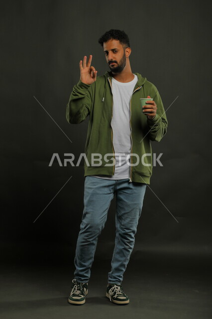 الإعجاب والاستمتاع بتناول المشروبات الساخنة، منتج محلي وطني، الحيوية والنشاط عند شرب الكافيين، بورتريه لرجل عربي سعودي يرتدي الزي الكاجوال يحمل بيده كوب ورقي بإيماءات علامة جودة القهوة السعودية، صورة الجسم بالكامل، خليفية سوداء