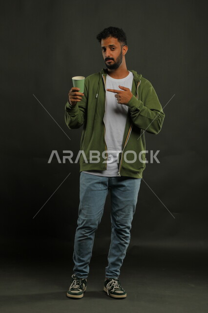 احتساء المشروبات الساخنه والأعشاب الشتوية الدافئة، منتج محلي وطني، الاستمتاع بتناول القهوة العربية، تعديل المزاج بتناول الكافيين، بورتريه لرجل عربي خليجي سعودي يرتدي الزي الكاجوال يشير بيده إلى كوب من الورق المقوى, صورة الجسم بالكامل، خلفية سوداء