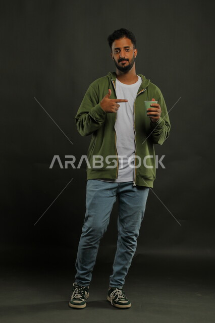 احتساء المشروبات الساخنه والأعشاب الشتوية الدافئة، منتج محلي وطني، الاستمتاع بتناول القهوة العربية، تعديل المزاج بتناول الكافيين، بورتريه لرجل عربي خليجي سعودي يرتدي الزي الكاجوال يشير بيده إلى كوب من الورق المقوى, صورة الجسم بالكامل، خلفية سوداء