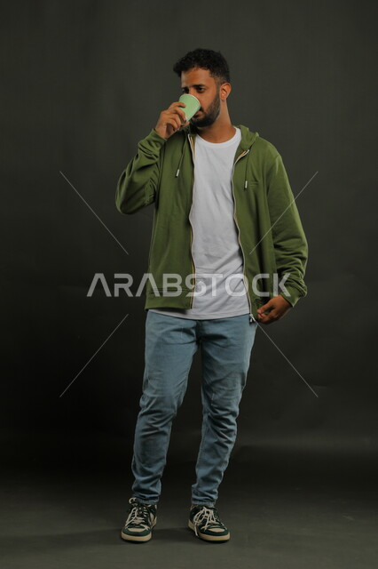 تعديل المزاج بتناول الكافيين، احتساء المشروبات الساخنه والأعشاب الشتوية الدافئة، منتج محلي وطني، بورتريه لرجل عربي خليجي سعودي يرتدي الزي الكاجوال يشرب القهوة العربية بإيماءات الاستمتاع, صورة الجسم بالكامل، خلفية سوداء