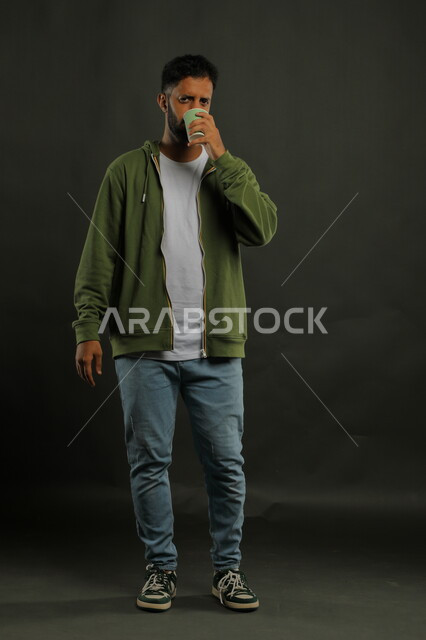 تعديل المزاج بتناول الكافيين، احتساء المشروبات الساخنه والأعشاب الشتوية الدافئة، منتج محلي وطني، بورتريه لرجل عربي خليجي سعودي يرتدي الزي الكاجوال يشرب القهوة العربية بإيماءات الاستمتاع, صورة الجسم بالكامل، خلفية سوداء