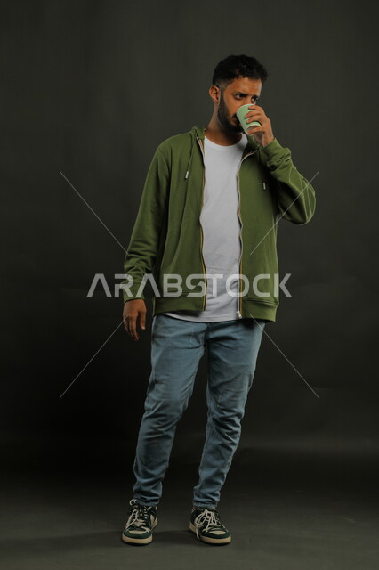 تعديل المزاج بتناول الكافيين، احتساء المشروبات الساخنه والأعشاب الشتوية الدافئة، منتج محلي وطني، بورتريه لرجل عربي خليجي سعودي يرتدي الزي الكاجوال يشرب القهوة العربية بإيماءات الاستمتاع, صورة الجسم بالكامل، خلفية سوداء