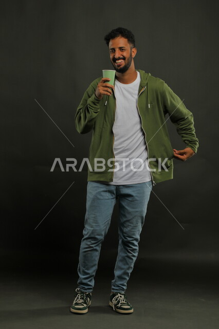 الاستمتاع بشرب القهوة، تعديل المزاج بتناول الكافيين، احتساء المشروبات الساخنه والأعشاب الشتوية الدافئة، منتج محلي وطني، بورتريه لرجل عربي خليجي سعودي يرتدي الزي الكاجوال يمسك بيده كوب من الورق المقوى بإيماءات السعادة والفرح, صورة الجسم بالكامل، خلفية سوداء