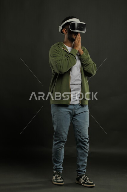 Watching favorite movies with enthusiasm, simulating the imaginary world, portrait of a Saudi Gulf Arab young man wearing casual clothes and 3D VR glasses, placing his palms over his mouth with waiting gestures, interacting and merging with virtual reality games, full body image, black background.