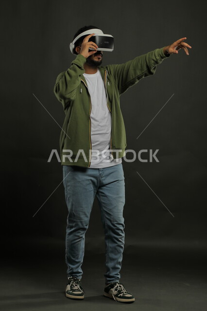 التقنيات المتطورة الحديثة، استخدام حاسة اللمس للنقر على عنصر ما, بورتريه لرجل عربي خليجي سعودي يرتدي الزي الكاجوال ونظارة الواقع الافتراضي ثلاثية الأبعاد يشير إلى شيء ما بأصبع السبابة, محاكاة العالم الخيالي, صورة للجسم بالكامل, خلفية سوداء
