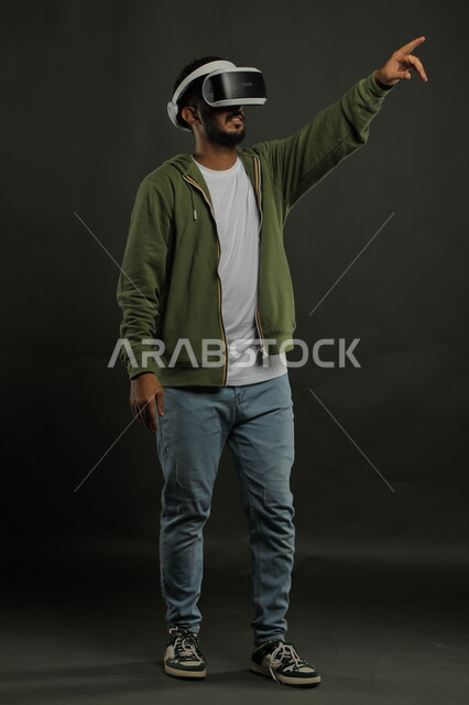 التقنيات الحديثة المتطورة، استخدام حاسة اللمس للنقر على عنصر ما, بورتريه لرجل عربي خليجي سعودي يرتدي الزي الكاجوال ونظارة الواقع الافتراضي ثلاثية الأبعاد يشير إلى شيء ما بأصبع السبابة, محاكاة العالم الخيالي, صورة للجسم بالكامل, خلفية سوداء