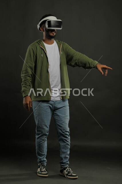 الاشارة الى شيء ما ،بورتريه لرجل عربي خليجي سعودي يرتدي الزي الكاجوال ونظارة الواقع الافتراضي ثلاثية الأبعاد يقوم باستخدام حاسة اللمس للنقر على عنصر ما, محاكاة العالم الخيالي, صورة للجسم بالكامل, خلفية سوداء