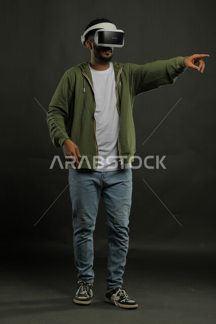 استخدام حاسة اللمس للنقر على عنصر ما , بورتريه لرجل عربي خليجي سعودي يرتدي الزي الكاجوال ونظارة الواقع الافتراضي ثلاثية الأبعاد يشير إلى شيء ما بأصبع السبابة, محاكاة العالم الخيالي, التقنيات الحديثة المتطورة، صورة للجسم بالكامل, خلفية سوداء 