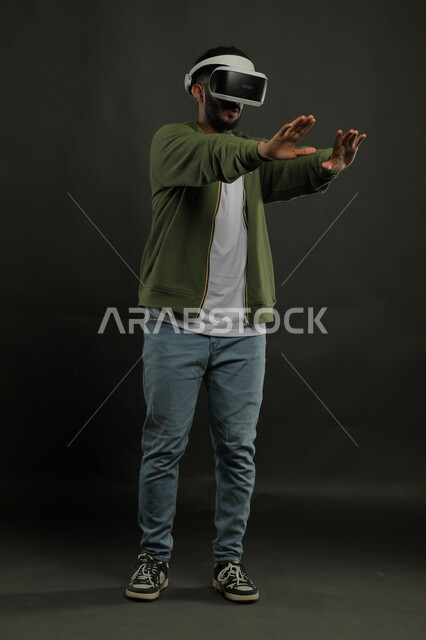 محاولة لمس عنصر ما ومحاكاة العالم الخيالي , إيماءات الدهشة والاستمتاع, بورتريه لرجل عربي سعودي خليجي يرتدي الزي الكاجوال ونظارة في آر ثلاثية الأبعاد, التفاعل والاندماج مع ألعاب الواقع الافتراضي, تقنيات وأجهزة متطورة, صورة الجسم بالطول الكامل, خلفية سوداء