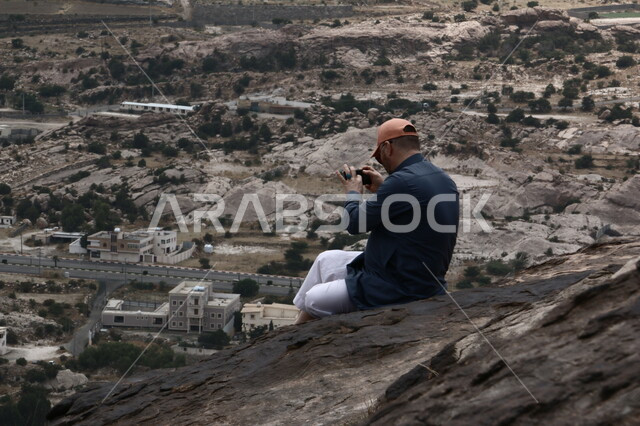 التكوينات الصخرية في القمم والمرتفعات الجبلية, الرحلات السياحية في جبال تنومة في منطقة عسير, سلسلة جبال السروات, المناظر الطبيعة والاستمتاع بقضاء وقت ممتع في المعالم والمناطق العالية, صورة من الخلف لزائر يقوم بالتقاط صور للطبيعة في المملكة العربية السعودية