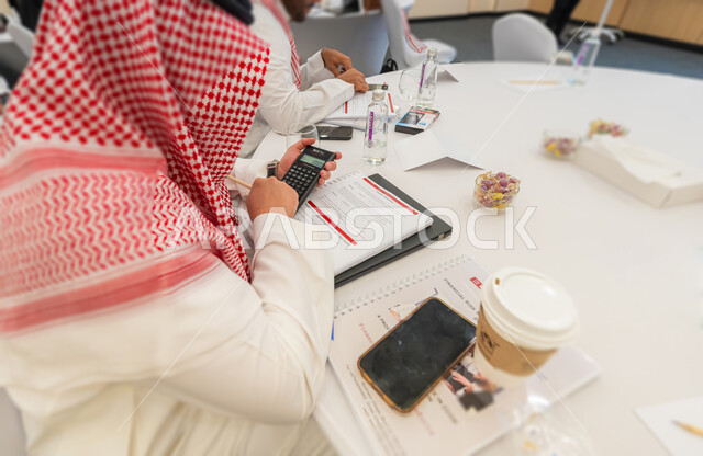 مهن ووظائف وأعمال مكتبية سعودية, استخدام الآلة الحاسبة لإجراء العمليات الحسابية, استراتيجية العمل على إدراة الحسابات وتدوين الملاحظات, صورة مقربة من الخلف لرجل عربي خليجي سعودي يرتدي الثوب التقليدي والشماغ يقوم بالإحصائيات الرقمية