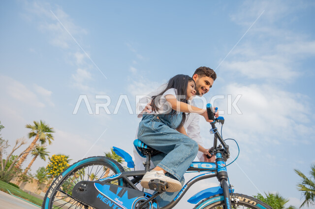 ممارسة رياضة ركوب الدراجات الهوائيه, فتاة عربية خليجية سعودية تقوم بقيادة الدراجة بمساعدة شقيقها في إحدى الحدائق العامة, أنشطة ترفيهيه مسلية, قضاء اوقات عائلية ممتعة في وقت العطلة الصيفية, ايماءات الحماس والسرور، منتجعات وشاليهات راقية في السعودية