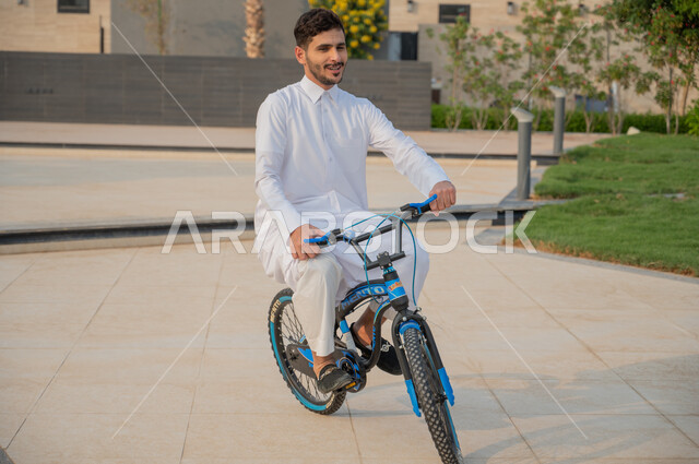أنشطة بدنية ترفيهيه مسلية, ممارسة هواية ركوب الدراجات الهوائيه, شاب عربي خليجي سعودي يرتدي الثوب التقليدي يقوم بقيادة الدراجة في إحدى الحدائق العامة,  ايماءات الحماس والسرور، منتجعات وشاليهات راقية في السعودية