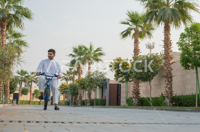 أنشطة بدنية ترفيهيه مسلية, ممارسة هواية ركوب الدراجات الهوائيه, شاب عربي خليجي سعودي يرتدي الثوب التقليدي يقوم بقيادة الدراجة في إحدى الحدائق العامة,  ايماءات الحماس والسرور، منتجعات وشاليهات راقية في السعودية