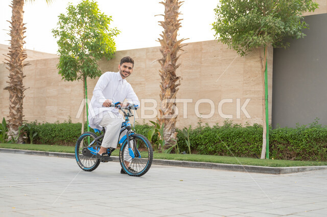 منتجعات وشاليهات راقية في السعودية، ممارسة هواية ركوب الدراجات الهوائيه, شاب عربي خليجي سعودي يرتدي الثوب التقليدي يقوم بقيادة الدراجة في إحدى الحدائق العامة, ايماءات الحماس والسرور، أنشطة بدنية ترفيهيه مسلية