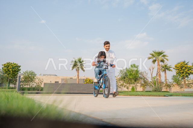 المشاركة مع الأبناء في الأنشطة الترفيهية، تعليم الأولاد التوازن واللعب في الدراجات الهوائية، رجل عربي خليجي سعودي يرتدي الزي التقليدي يساعد إبنته في قيادة الدراجة في إحدى الحدائق العامة، إيماءات الفرح في قضاء أوقات عائلية ممتعة، الطبيعة الخضراء في المملكة.