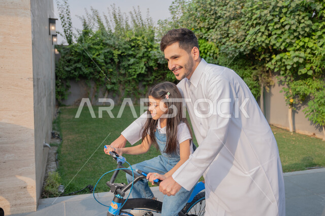 قضاء وقت ممتع مع الأبناء في الأنشطة الترفيهية، استراحات ومنتزهات عائلية، أب عربي خليجي سعودي يرتدي الزي التقليدي يساعد ابنته في ركوب الدرجة بإيماءات الفرح، تعليم الأولاد قيادة الدراجة الهوائية، الطبيعة الخضراء في المملكة العربية السعودية .
