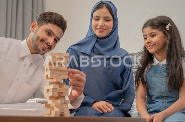 أنشطة ترفيهية لتنمية مهارات التفكير والذكاء عند الأطفال، قضاء إجازة ممتعة في اللعب مع الأبناء، عائلة عربية خليجية سعودية مبتسمة تلعب لعبة جينغا بإيماءات الفرح، رص الألواح الخشبية، ألعاب التحدي المسلية.