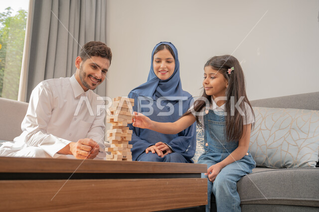 مهارة رص الألواح الخشبية، قضاء إجازة ممتعة في اللعب مع الأبناء، عائلة عربية خليجية سعودية تجلس في غرفة المعيشة تلعب لعبة جينغا بإيماءات الفرح، أنشطة ترفيهية لتنمية مهارات التفكير والذكاء عند الأطفال، ألعاب عائلية مسلية.
