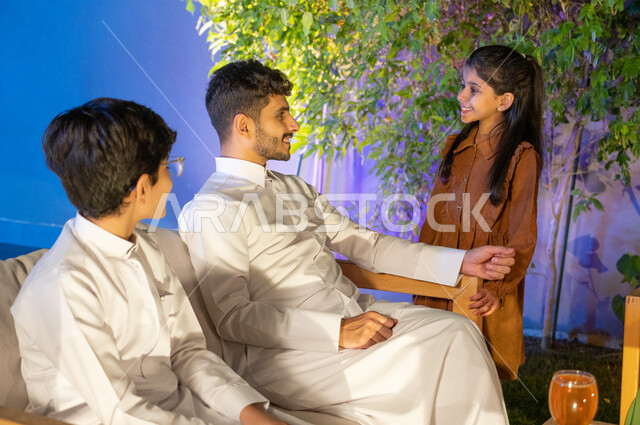 تبادل النظرات والاحاديث بين الفتاة ووالدها، اجواء اسرية مليئة بالسعادة و السرور، أب عربي خليجي سعودي يرتدي الثوب التقليدي يجلس على اريكة مريحة بجانب ولده في احدى الشاليهات الراقية، قضاء اوقات عائلية ممتعة