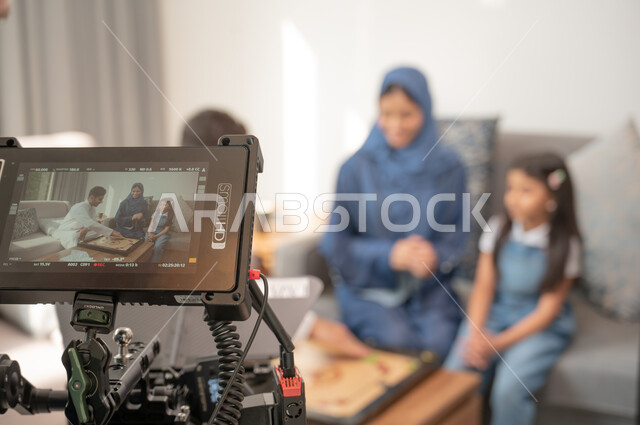 لقطة مقربة لكاميرا حديثة ومتطورة، إيماءات الاندماج بالتسلية والترفيه، خلف الكواليس لجلسة تصوير في غرفة المعيشة لرجل عربي خليجي سعودي يرتدي الزي التقليدي يلعب مع أسرته لعبة الكيرم، قضاء إجازة ممتعة في اللعب مع الأبناء بألعاب التحدي المسلية.