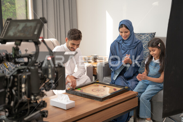 إيماءات الاندماج بالتسلية والترفيه، خلف الكواليس لجلسة تصوير في غرفة المعيشة لرجل عربي خليجي سعودي يرتدي الزي التقليدي يلعب مع أسرته لعبة الكيرم، لقطة مقربة لكاميرا حديثة ومتطورة، قضاء إجازة ممتعة في اللعب مع الأبناء بألعاب التحدي المسلية.