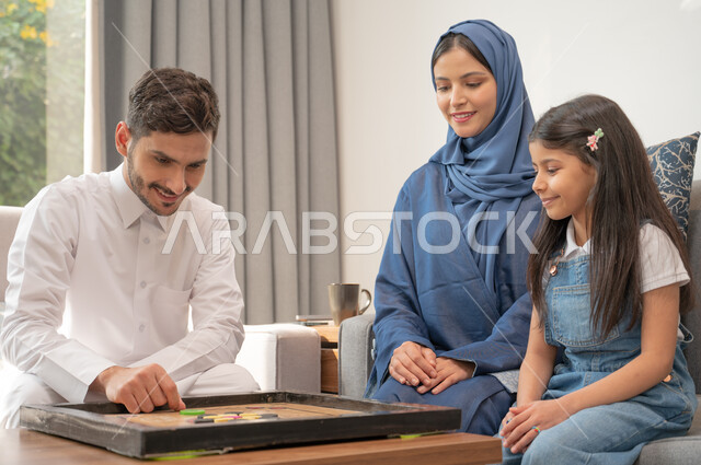 قضاء أوقات ممتعة في اللعب مع الأبناء، صورة مقربة لعائلة عربية خليجية سعودية تجلس في غرفة المعيشة تلعب لعبة الكيرم بإيماءات المرح، أنشطة ترفيهية لتنمية مهارات التفكير والذكاء عند الأطفال، ألعاب عائلية مسلية.