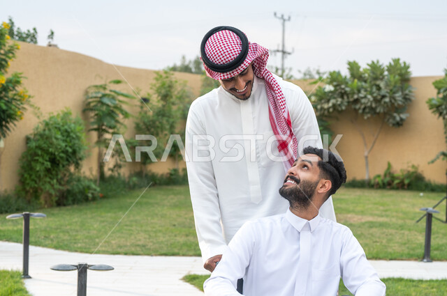 التسامر والضحك مع المرضى، العطف والحنان بين أفراد الأسرة، أخ سعودي يقوم بالاهتمام بالجانب النفسي والجسدي لشقيقه, صورة مقربة لشاب عربي سعودي خليجي من ذوي الهمم يرتدي الزي التقليدي يجلس على كرسي متحرك يتبادل الاحاديث الجانبيه, قضاء الوقت في المنتزه مع العائلة